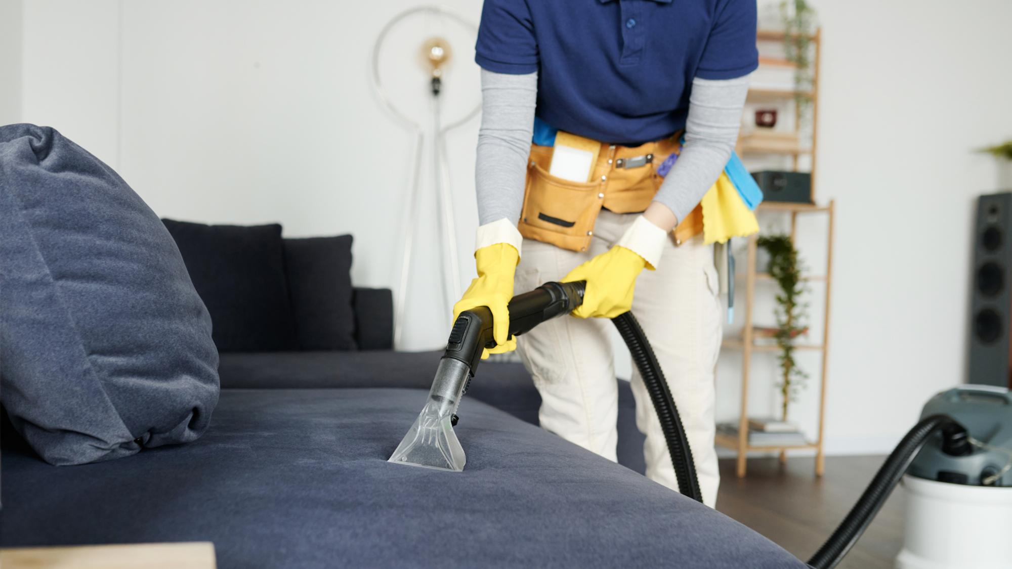 A person cleaning a dark blue sofa using a vacuum cleaner with a hose attachment, wearing yellow gloves and a tool belt, in a well-lit room with plants and modern furniture.